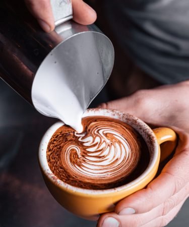 A barrister's hands are seen pouring frothed milk into a large coffee cup, forming a squiggly pattern on the surface of the coffee.